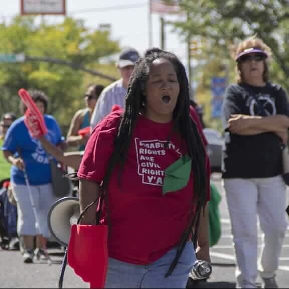 Anita marching and chanting with ADAPT members behind her. She is wearing a red shirt with image of state of Arkansas that reads disability rights are civil rights y'all.