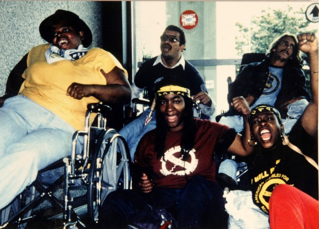 Paulette Patterson in a manual wheelchair, Anita Cameron and another woman protester sit on the floor mouths open, all chanting in unison in Atlanta 1989. Anita and the other woman wear ADAPT headbands and have their fists raised in the power sign. Behind them George Roberts and Claude Holcomb are in their wheelchairs up against a a glass door that reads "DO NOT ENTER (with and ADAPT sticker over the center)" and "OUT." George is also chanting, Claude looks off to the side. Photo by Tom Olin.