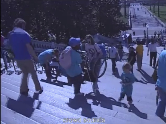 Anita, a Black person wearing blue jeans, blue T-shirt, blue baseball cap, red bandanna and a pink backpack, is walking backwards up the steps of the US Capitol. She is holding a white tactile cane. She is helping another person carry a manual wheelchair. They are surrounded by other people, including a young child, who might be either Caitlin or Lincoln Blank.