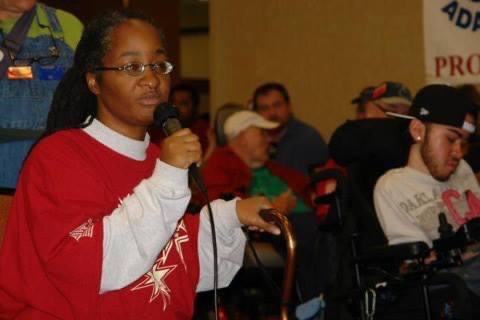 Anita a black woman with dreadlocks wearing glasses, a red ADAPT t-shirt over a longsleeve white shirt holding a microphone and cane addressing a crowd of ADAPT members in a room.