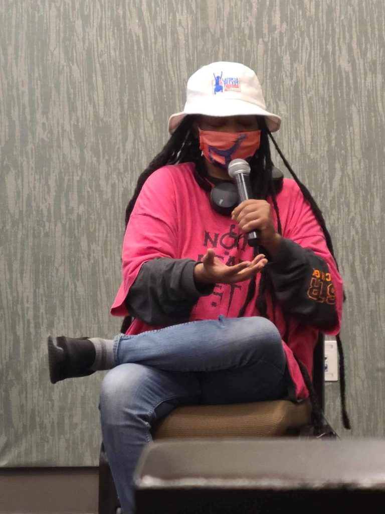 Anita speaking at the 2023 NYAIL conference on how communities of color can organize and fight against assisted suicide laws. She is wearing a mask, pink Not Dead Yet shirt, white LaTonya Reeves Act hat, and jeans sitting in a chair holding a microphone.