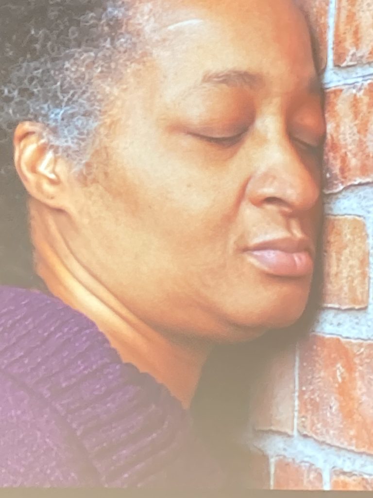 Anita in a purple sweater with her eyes closed leaning against a brick wall.