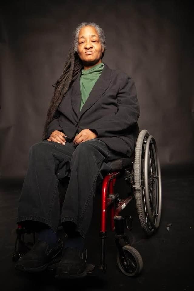 Anita Cameron sits proudly in her wheelchair against a dark background. They are wearing black pants and blazer and a green turtleneck.
