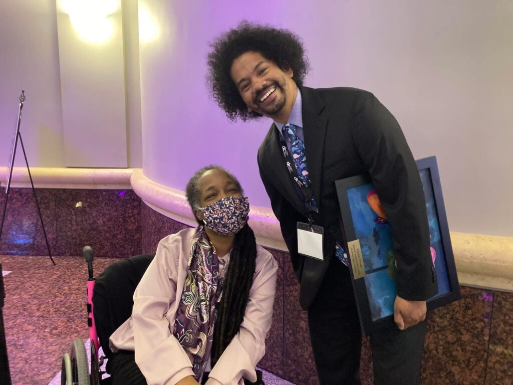 Anita a black woman with dreadlocs and some gray hair sitting in her hot pin wheelchair wearing a floral print mask, pink jacket, and plum and other warm toned print blouse. She is posing next to Mychal Threets a black and Mexican with an afro wearing a black suit, purple shirt and blue tie with a print on it. He is holding an award plaque from Disability Rights California. Both are smiling in a banquet hall.