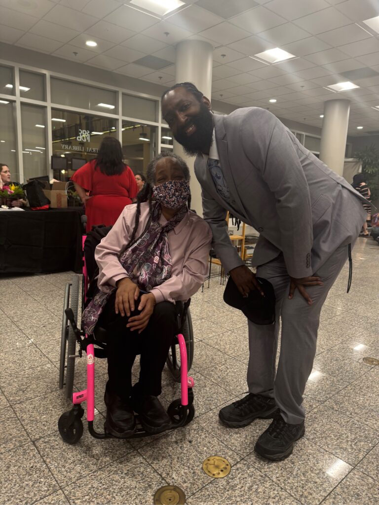 Anita a black woman with dreadlocs and some gray hair sitting in her hot pin wheelchair wearing a floral print mask, pink jacket, and plum and other warm toned print blouse. She is posing next to Antoine Hunter, a deaf artist and choreographer with his hair tied back and a long beard. He is bending down towards Anita and he's wearing a light gray suit, white shirt and a print designed tie. They are in a hallway at the Disability Rights California Gala.