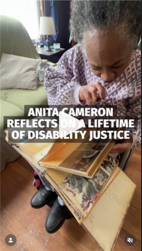 Screenshot of WXXI/Rochester NY NPR Instagram Reel of interview with Anita. Text reads Anita Cameron reflects on a lifetime of disability justice. Anita, a blac woman with dreadlocs and gray hair, is sitting next to a mint green couch in her hot pink wheelchair wearing a purple and gray patterned knit sweater looking through an old photo album of hers.