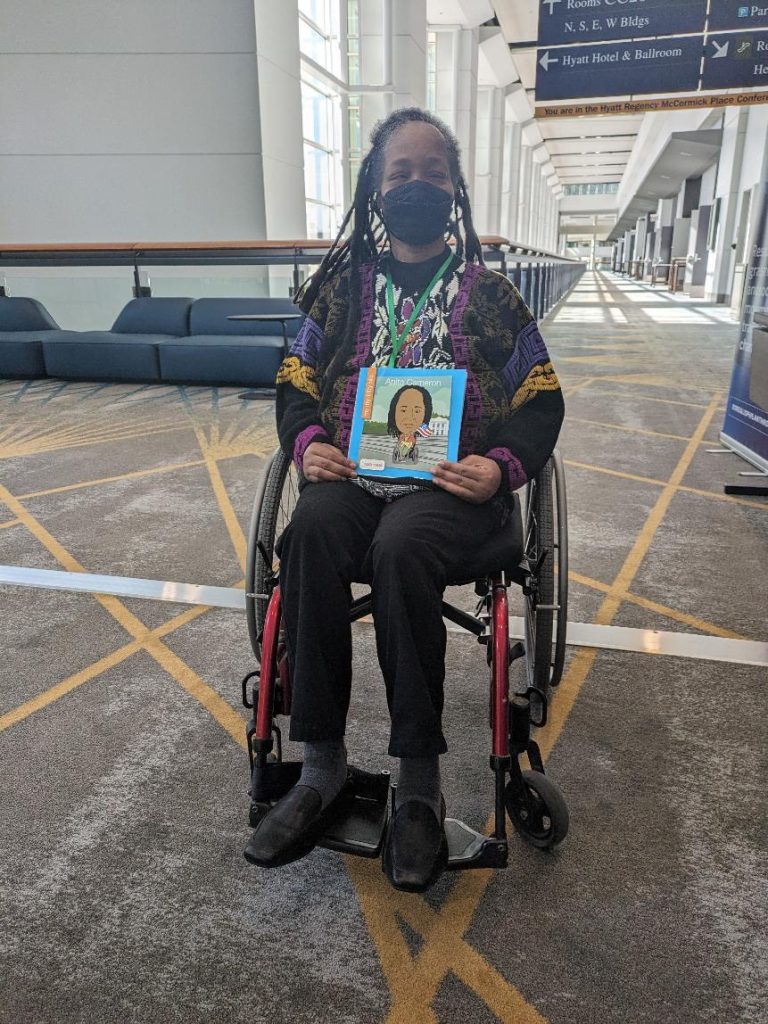 Anita Cameron holds a copy of their book, My Itty Bitty Bio. Anita is wearing a mask and in a wheelchair inside a hotel lobby at a conference. 