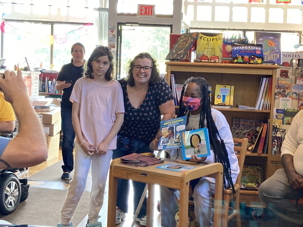 Anita wearing a mask sitting at a table holding an event poster and My Itty Bitty Bio Book with an adult and child standing next to her smiling for a camera.